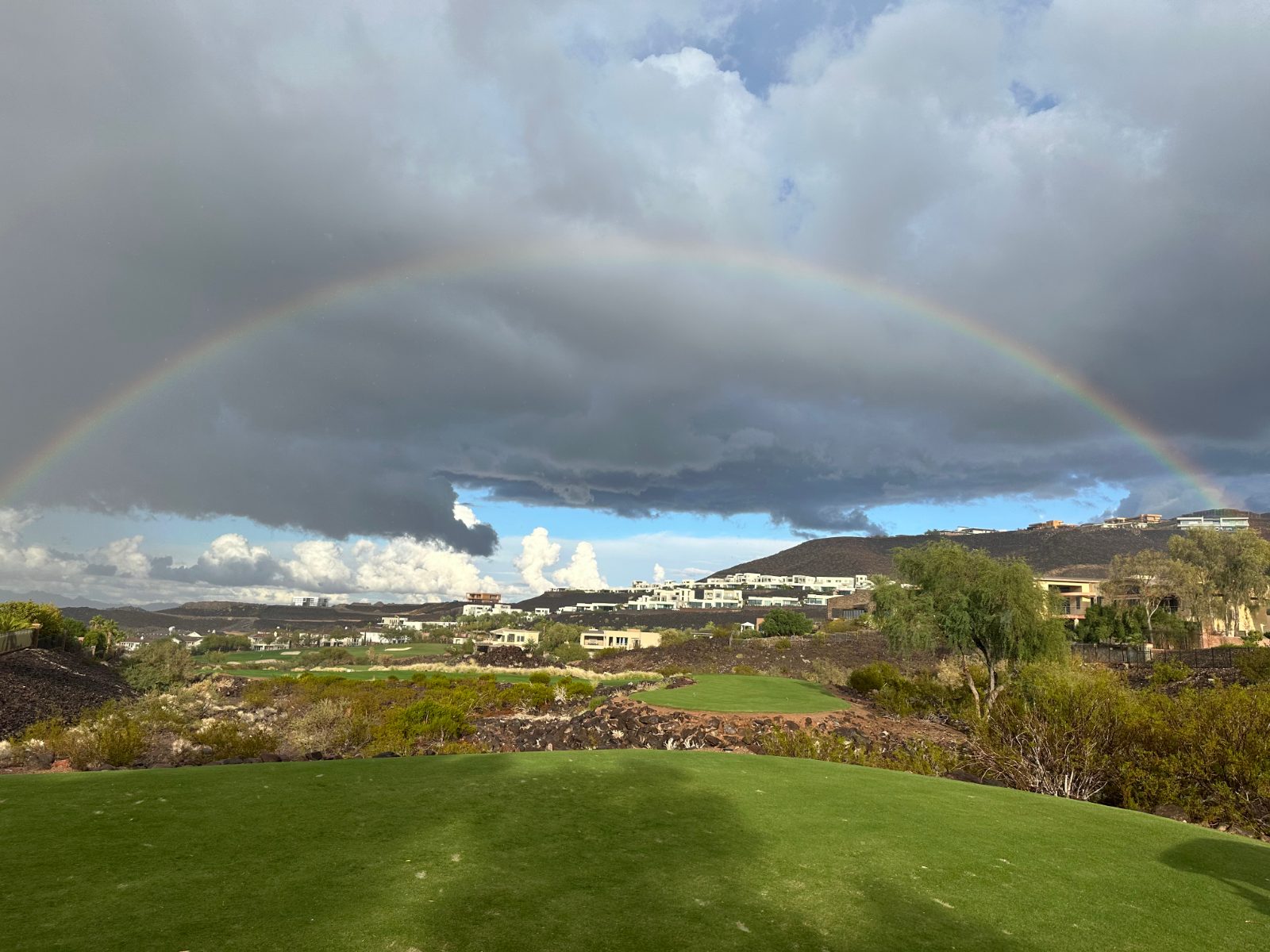 A full rainbow arcs across dark gray storm clouds above a green golf fairway, with desert shrubs, trees, and hillside homes in the distance under patches of blue sky.