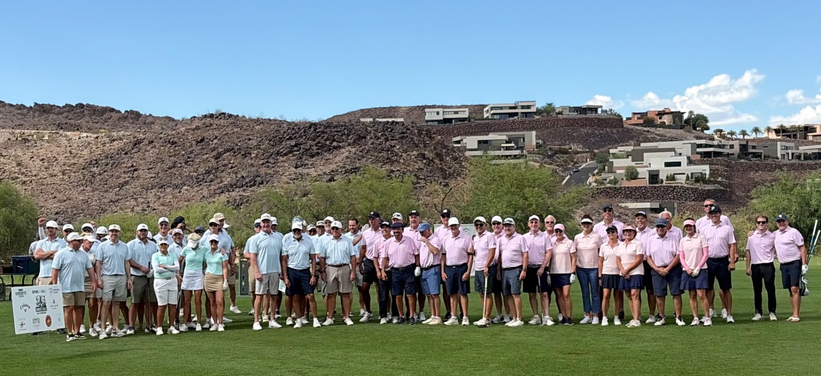 A large group of golfers poses on a fairway in two teams, one in light blue shirts and one in pink, with rocky desert hills and modern homes behind under a blue sky.